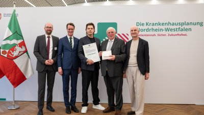 MP Wüst und Minister Lauman posieren mit Förderbescheidempfängern und eine Urkunde für ein Foto