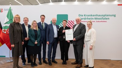 MP Wüst und Minister Lauman posieren mit Förderbescheidempfängern und eine Urkunde für ein Foto