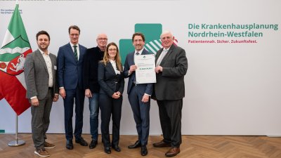 MP Wüst und Minister Lauman posieren mit Förderbescheidempfängern und eine Urkunde für ein Foto