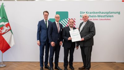 MP Wüst und Minister Lauman posieren mit Förderbescheidempfängern und eine Urkunde für ein Foto
