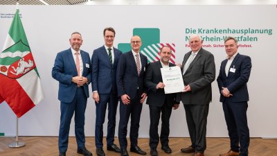 MP Wüst und Minister Lauman posieren mit Förderbescheidempfängern und eine Urkunde für ein Foto