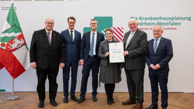 MP Wüst und Minister Lauman posieren mit Förderbescheidempfängern und eine Urkunde für ein Foto