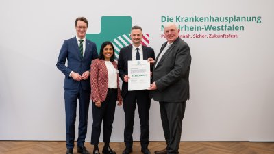 MP Wüst und Minister Lauman posieren mit Förderbescheidempfängern und eine Urkunde für ein Foto