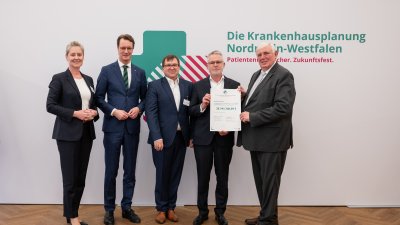 MP Wüst und Minister Lauman posieren mit Förderbescheidempfängern und eine Urkunde für ein Foto