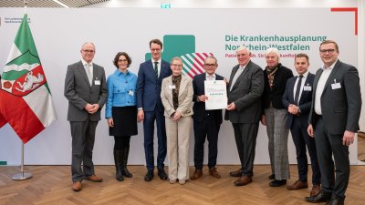 MP Wüst und Minister Lauman posieren mit Förderbescheidempfängern und eine Urkunde für ein Foto