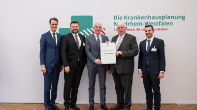 MP Wüst und Minister Lauman posieren mit Förderbescheidempfängern und eine Urkunde für ein Foto