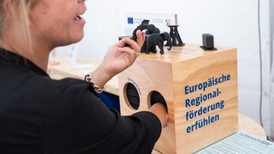 Wirtschaftsministerin Mona Neubaur fasst in eine Holzbox mit dem Schriftzug "Europäische Regionalförderung erfühlen"