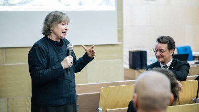 Eine vortragende Person erzählt etwas im Hörsaal, rechts sitzt MP Wüst
