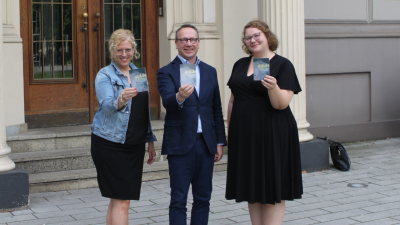 Zwei Frauen und ein Mann stehen vor einer Eingangstür und halten ein Ratgeberheft hoch