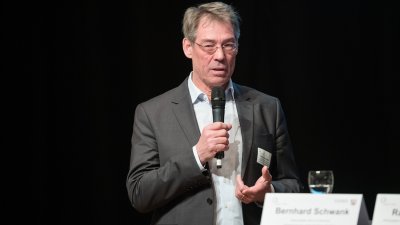 Ein Redner im grauen Anzug und weißem Hemd mit Mikrofon in der Hand am Rednerpult.