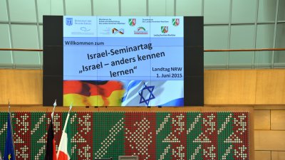 Über dem rednerpult hängt ein Monitor mit dem Titel der Veranstaltung "Israel-Seminartag. Isreal anders kennenlernen"
