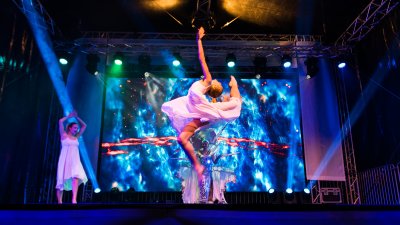Showauftritt von 3 Tänzerinnen. Mitig springt eine Ballerina in die Höhe, wobie sie ihr linkes Bein hinten hochzieht und gleichzeitig ihren Rücken nachhinten wölbt und den linken Arm nach oben gestreckt hat.