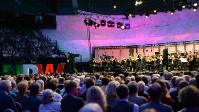 Der Dirigent steht auf der Bühne, im Hintergrund das Orchester, im Vordergrund Köpfe des Publikums