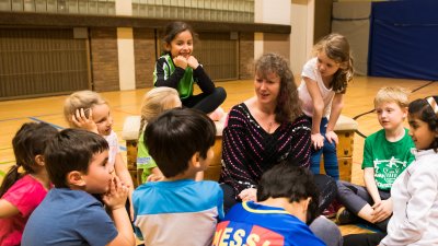 Staatssekretärin Andrea Milz sitzt umringt von Grundschulkindern in einer Sporthalle. IN ihrer Hand hält sie ein Pixi-Buch.