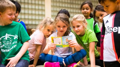 Eine Gruppe vo Grundschulkindern schauen sich ein Pixi-Buch an. Im Hintergrund sieht man die Glaskachelwand der Sporthalle.