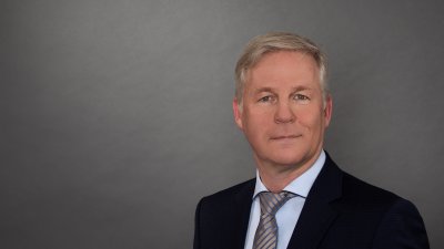 Ein Mann mit hellen Haaren, blauem Jackett, hellblauem Hemd und grau-blau gestreifter Kravatte