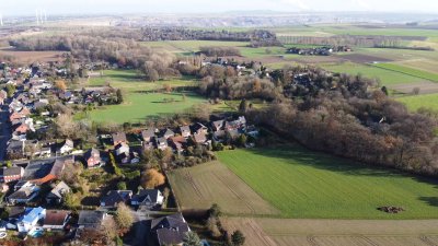 Aus der Vogelperspektive sieht man auf der linken Seite einige Häuser, die an grüne Felder und ein Waldstück grenzen