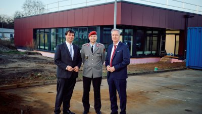 zwei Männer mit Anzug und ein Mann in Bundeswehr-Uniform stehen vor einem roten Gebäude