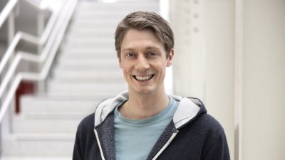 Ein Mann mit hellblauem Tshirt und dunkelblauer Kapuzenjacke steht lächelnd vor einem Treppenaufgang