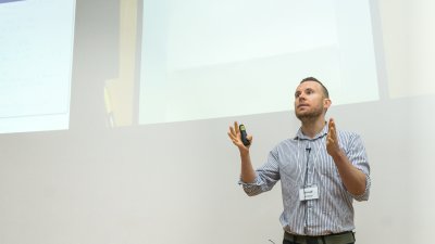 Ein Mann in hellblauem Hemd steht gestikulierend vor einer Leinwand 
