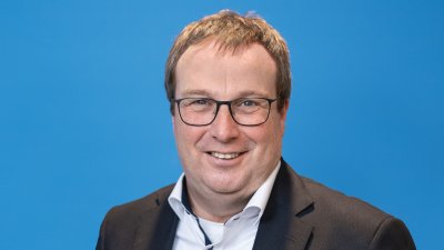 Ein Mann mit braunen kurzen Haaren, Brille, weißem Hemd und schwarzem Jackett lächelt in die Kamera