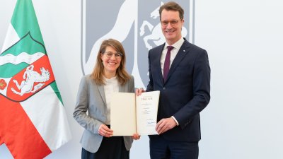 Rechts neben der NRW-Fahne steht Verena Schäffer mit langen blonden Haaren und Brille im grauen Sakko und weißem Hemd lächelnd neben MP Wüst im schwarzen Anzug mit roter Krawatte