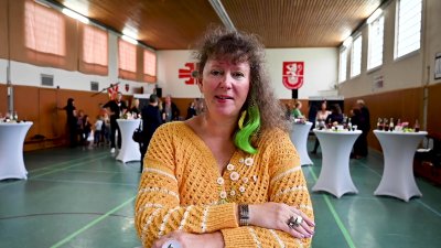 Staatssekretärin MIlz im orangefarbenen  mit weißen Applikationen in einer Sporthalle. Im Hintergrund sieht man festlich dekorierte Stehtische, die in weißen Tischtüschern eingekleidet sind.