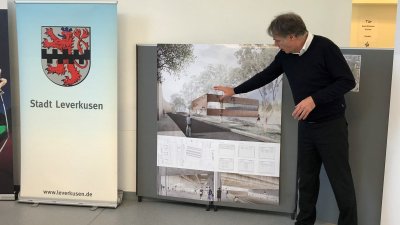 Rechts steht ein Herr im weißen Hemd, schwarzen Pullover und schwarzer Hose auf ein Bild zeigend, dabei hält er einen Vortrag. Links sieht man das Wappen von der Stadt Leverkusen mit rotem Löwen auf weißen Grund und einem schwarzen Balkenmuster mittig quer verlaufend.
