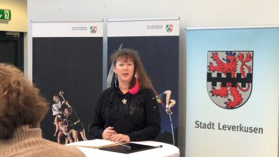 Staatssekretärin Andrea Milz sitzt an einem Tisch, vor ihr sitzt ein Publikum. Rechts sieht man das Wappen von Leverkusen, den roten Löwen auf weißen Grund und einem schwarzen Balkenmuster mittig quer verlaufend.