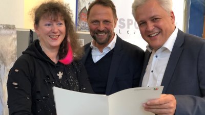 Staatssekretärin Milz steht links neben zwei Herren von der Stadt Leverkusen, gemeinsam halten sie den Zuwendungsbescheid.