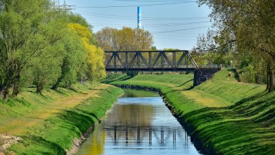 Blick auf einen begrünten Kanal in Oberhausen mit einer Stahlbrücke, spiegelndem Wasser und Bäumen entlang der Ufer. Im Hintergrund sind Stromleitungen und ein markanter Industrieschornstein zu sehen