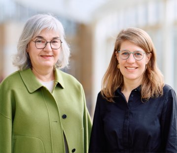 Petra Ladenburger, eine ältere Frau mit grauen schulterlangen Haaren und einer runden Brill in grünem Kostüm steht links neben Verena Schäffer in einem schwarzen Hemd