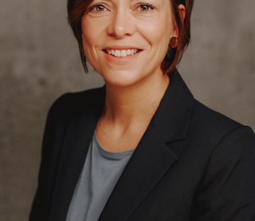 Eine Frau mit kurzen braunen Haaren, braunen Augen, in schwarzem Blazer lächelt in die Kamera