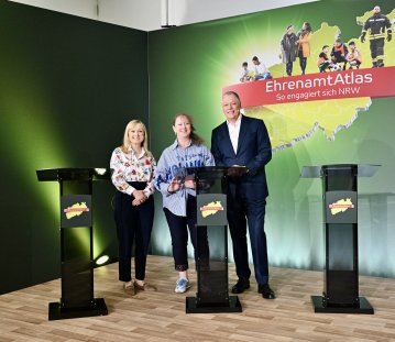 zwei Frauen und ein Mann stehen hinter drei Pulten vor einer grünen Leinwand mit einem EhrenamtAtlas Schriftzug