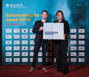 Zwei Personen präsentieren gemeinsam den „Senkrechtstarter Award 2026“ vor einer Sponsorenwand mit Logos und der Aufschrift „BOCHUM Wirtschaftsentwicklung“. Rechts ist Ministerin Mona Neubaur zu sehen.