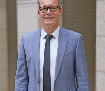 Ingo Dudenhausen steht im blauen Anzug und dunkelblauer Krawatt und weißem Hemd vor beigen Säulen. Er trägt eine Brille und lächelt frontal in die Kamera.