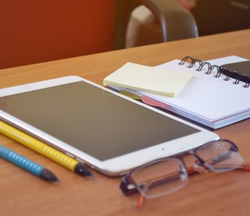 Auf einer Tischplatte liegen zwei Stifte, eine Brille, ein Tablet und ein Notizbuch