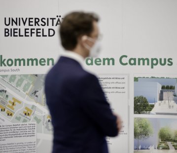 Ministerpräsident Wüst steht vor einem Informationsplakat der Universität Bielefeld