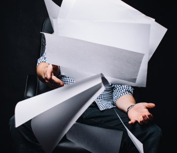 Ein Stapel weißes Papier, welches jemanden aus den Händen fällt. Der Hintergurnd ist schwarz.