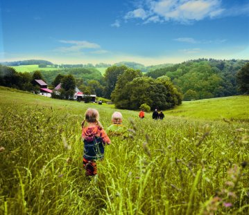 Eine Familie mit zwei Kindern streift durch eine hohe Wiese vor einem Dorf
