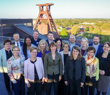 Gruppenfoto von etwa 15 Personen in formeller Kleidung auf einer Dachterrasse, die in die Kamera lächeln. Im Hintergrund steht ein großes Fördergerüst aus Stahl sowie Gebäude und eine grüne Landschaft. Die Personen stehen in zwei Reihen dicht beieinander.