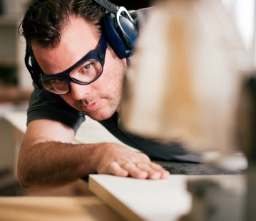Ein Mann im schwarzen T-Shirt mit schwarzer Brille und Kopfhörern als Lärmschutz sägt ein Holzbrett an einer Maschine.