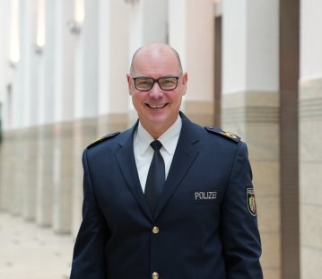Dirk Hulverscheidt, der neue Inspekteur der Polizei NRW steht in blauem Polizeianzug lächelnd in einer Halle. Auf dem Bild ist nur sein Oberkörper zu sehen mit dunkelblauem Sakko, weißem Hemd und blauer Krawatte. Er trägt eine Brille und hat sehr kurz geschnittene Haare.