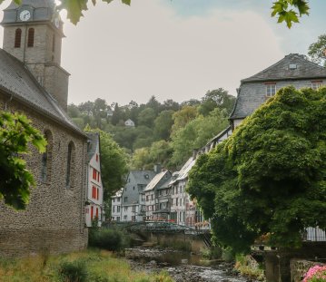 Historische Altstadt mit steinerner Kirche, Fachwerkhäusern und einem Bachlauf, eingerahmt von dichtem grünen Laub. Die idyllische Szene zeigt eine ruhige Flusslandschaft mit Brücke und blühenden Pflanzen.
