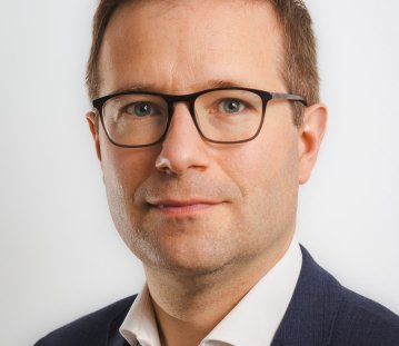 Ein Mann mit kurzen Haaren, Brille, weißen Hemd und blauem Jackett schaut in die Kamera