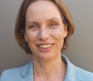Porträt einer lächelnden Frau mit kurzem, braunem Haar und freundlichem Blick. Sie trägt einen hellblauen Blazer über einem weißen Oberteil und blickt direkt in die Kamera. Der neutrale graue Hintergrund unterstreicht die professionelle und sympathische Ausstrahlung.