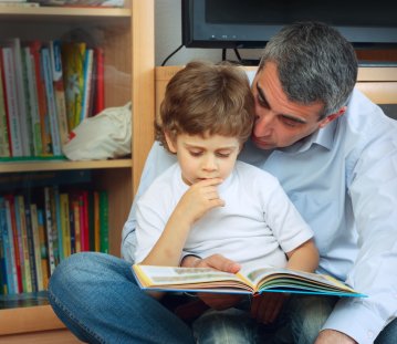 Ein Kind sitzt auf dem Schoß eines Mannes und sie lesen gemeinsam in einem Buch, im Hintergrund ist ein Bücherregal