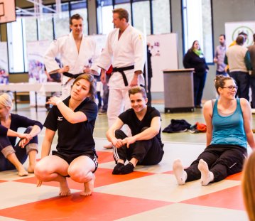 Mehrere Frauen bei Dehnübungen für einen Selbstverteidungskurs in einer Turnhalle. Auf dem Boden grüne und rote Matten im Schachbrettmuster. Im Hintergrund zwei Kampfsportler in weißen Anzügen mit schwarzen Gürteln.