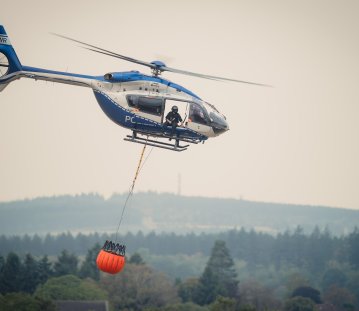 Zu sehen ist ein Löschhubschrauber der Polizei vor einem verrauchten Waldgebiet