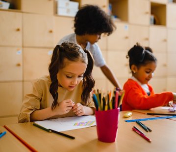 Drei Kinder sitzen an einem Holztisch und beschäftigen sich mit Zeichnen und Malen; im Vordergrund zeichnet ein Mädchen mit Zöpfen, während zwei weitere Kinder im Hintergrund kreativ arbeiten. Auf dem Tisch stehen ein roter Becher mit Buntstiften sowie verschiedene Stifte, Lineale und Zeichenmaterialien.